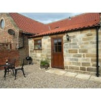 Stone cottage with red-tiled roof, wooden door, and gravel patio featuring a table, chairs, and a small grill.