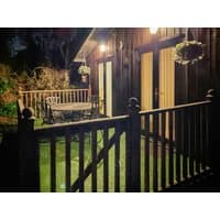 Wooden cottage exterior with patio, railing, hanging plants, and outdoor table set, warmly lit at night.