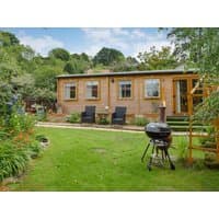 Wooden cottage with large windows, surrounded by lush garden, patio chairs, and a charcoal grill on green lawn.