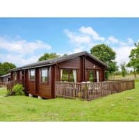 Brown wooden cottage with large windows, pitched roof, and wraparound deck, set in a grassy field with trees.