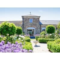Stone cottage with central tower, surrounded by lush gardens, trimmed hedges, and vibrant purple flowers.