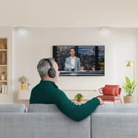 Black Hama 00221847 headphones with headband, used wirelessly by a person watching TV in a modern living room.