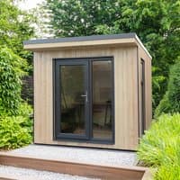 Insulated garden office with light wood paneling, black-framed glass doors, and flat roof, set in lush greenery.