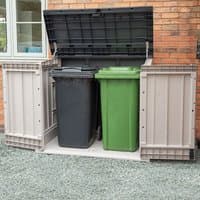 Grey Toomax 1200L plastic outdoor storage unit with open lid, showing space for two wheelie bins against a brick wall.