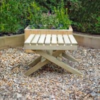 Forest slatted wooden garden table with crossed legs, natural finish, set on gravel with greenery in the background.