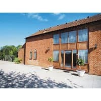 Red brick cottage with large wooden-framed windows, arched side windows, and potted plants on a sunny patio.