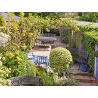 Garden patio with black metal benches, blue chairs, lush greenery, and a pergola, part of "Pax" by cottages.com.