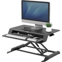 Black Fellowes sit-stand desk riser with adjustable height, supporting a monitor, keyboard, and small plant.