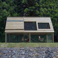 Wooden raised chicken coop with slanted roof, mesh windows, and enclosed run, set on grass with trees in the background.