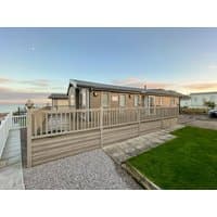 Beige wooden lodge with large windows, wraparound deck, and ocean view under a clear sky at sunset.