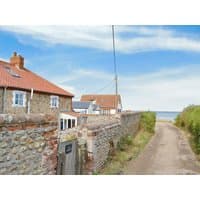 Stone cottage with red-tiled roof, chimney, and sea view, located along a rustic dirt path and stone wall.