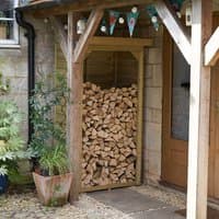 Wooden log store with slanted roof, filled with stacked firewood, placed under a porch next to a door.