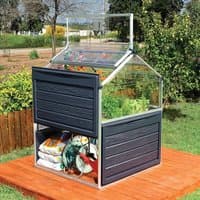 Small greenhouse with clear panels, black frame, and raised bed on a wooden deck, storing gardening supplies underneath.