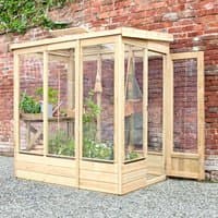 Wooden lean-to greenhouse with glazed back, clear panels, and door, set against a brick wall, housing green plants.