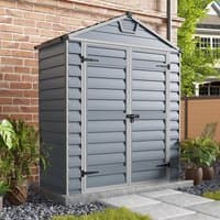 Grey plastic shed with skylight, double doors, and horizontal panel design, set against a brick wall with plants nearby.