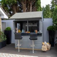 Gray shiplap garden bar with hinged serving hatch, two black stools, and a sloped roof, set on a wooden deck.
