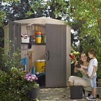 Beige and gray Keter Factor plastic garden shed with double doors, window, and sloped roof, surrounded by greenery.