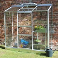 Greenhouse with clear panels, silver frame, sloped roof, and open vent, placed against a brick wall.