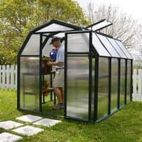 Green resin-framed greenhouse with clear panels, gabled roof, and open vent, set on grass near a white picket fence.