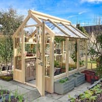 Wooden greenhouse with clear panels, pitched roof, and door, surrounded by plants and pots on a patio.