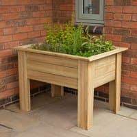 Wooden Forest Garden planter box with elevated legs, filled with herbs, placed against a brick wall.