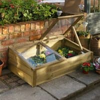 Large wooden cold frame with hinged transparent lid, housing colorful plants, set on a patio near a brick wall.