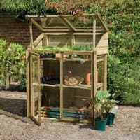 Wooden Forest Mini Greenhouse with glass panels, two shelves, and a sloped roof, housing plants and gardening tools.