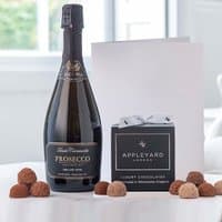 Prosecco Fidora bottle, black box of 12 handmade truffles, white occasion card, all on a white surface.