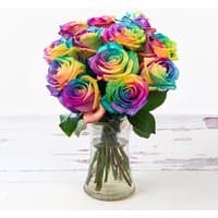 Bouquet of rainbow roses with vibrant multicolored petals in a clear glass vase on a white wooden surface.