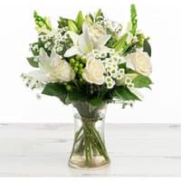 White rose and lily bouquet in a clear vase with greenery and small white flowers.